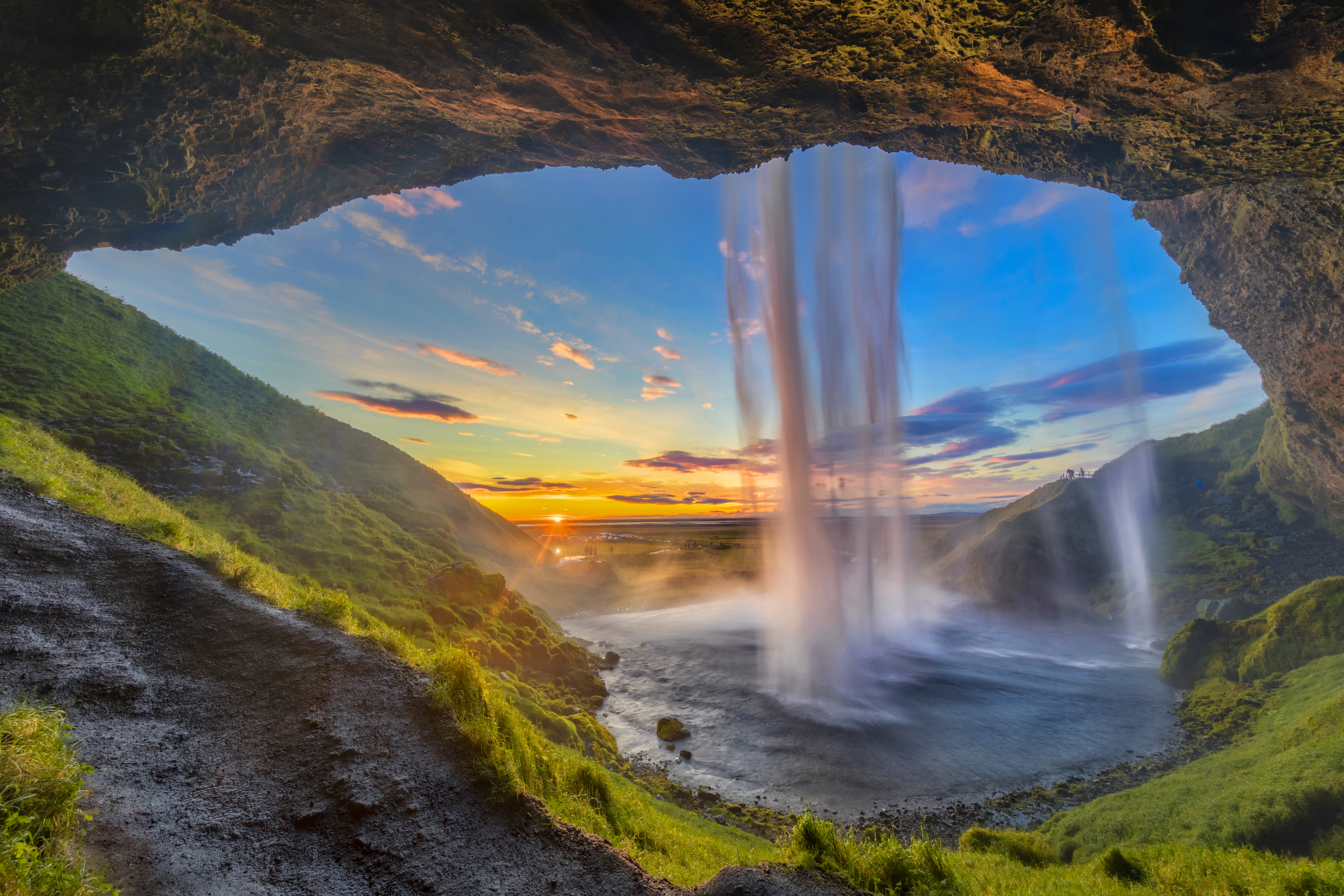 A view from behind Seljalandsfoss waterfall in Iceland, looking out from a cave at the vibrant sunset over the green landscape.