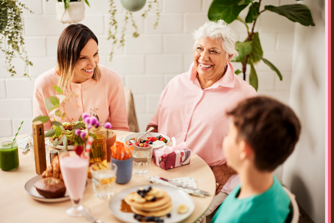 A colorful display of a mother, grandmother and child sitting at a table enjoying a meal together. Gifts and flowers, setting the scene for a special celebration. 