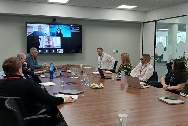 A group of people sitting around a long wooden boardroom table during a meeting, with three participants visible on a large video conferencing screen.