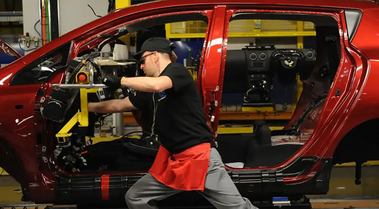 A factory worker is working on a bright red car body on an assembly line. The background shows factory environment.