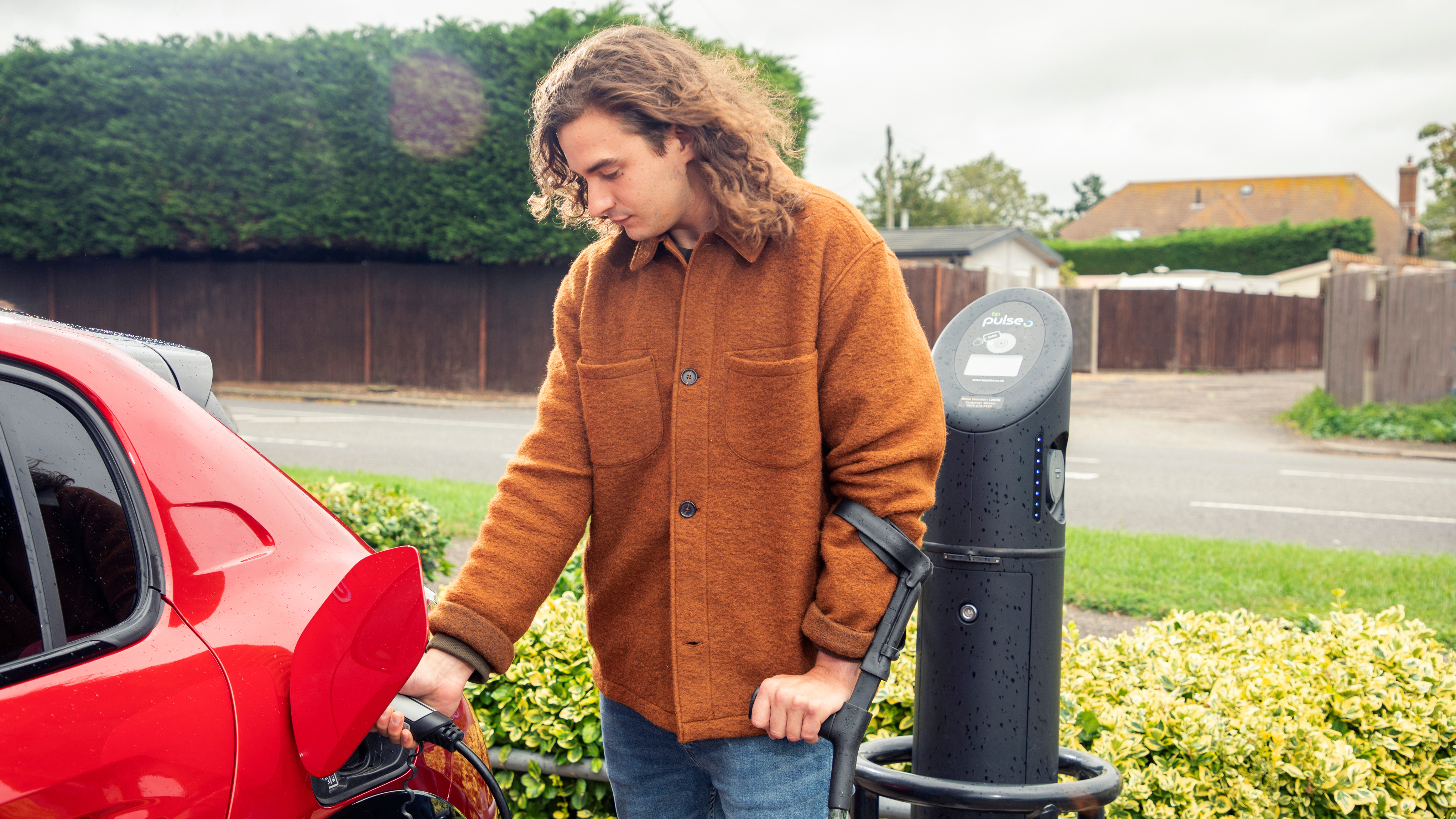 A man in a brown jacket charges a red electric car at a public charging point next to a road lined with hedges.