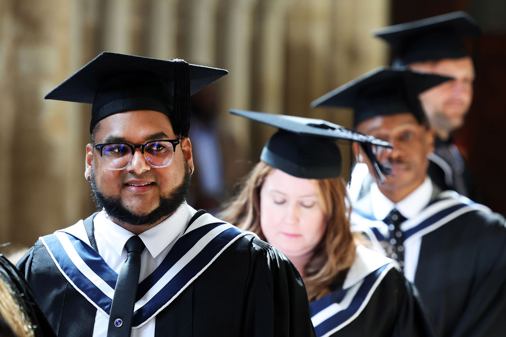 Close-up of a smiling graduate with a beard and glasses, walking in a procession with others behind