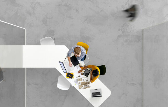 Top-down view of two co-workers sitting at a long table looking at laptops