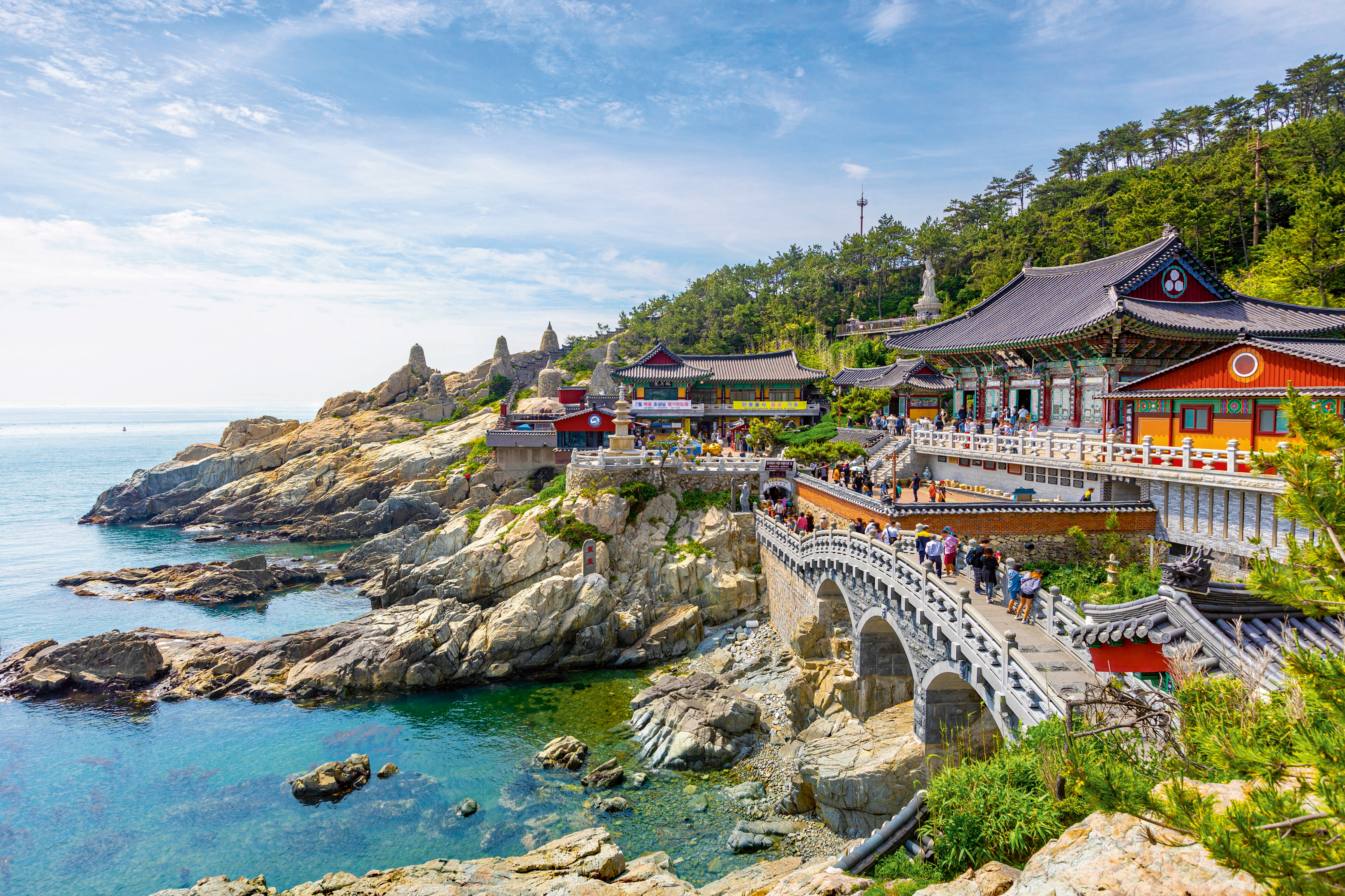 People cross a stone bridge toward traditional Korean buildings at the Floating Campus Busan, set against rocky cliffs and turquoise ocean waters.