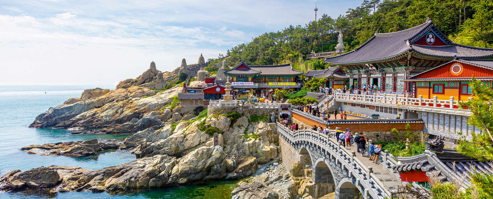 People cross a stone bridge toward traditional Korean buildings at the Floating Campus Busan, set against rocky cliffs and turquoise ocean waters.