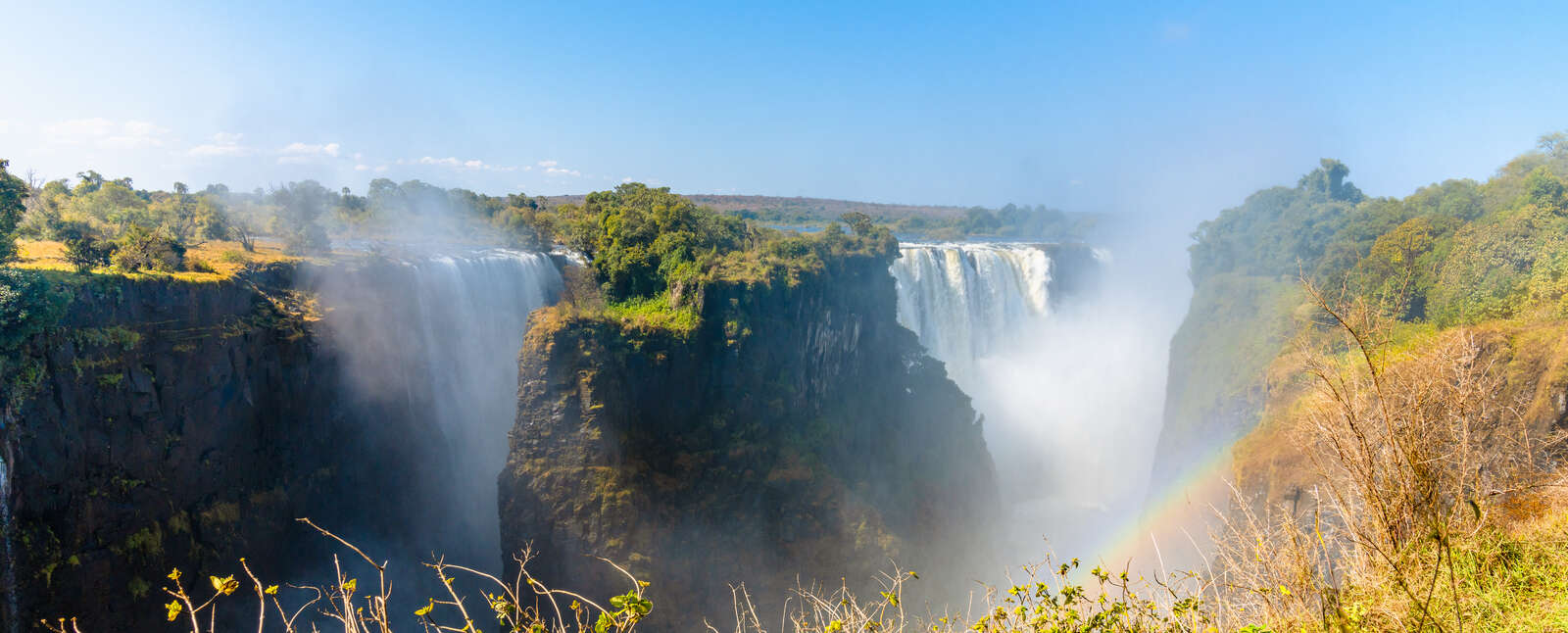 Powerful white water cascades over the cliffs of Victoria Falls in Zimbabwe, creating thick mist and a faint rainbow under a clear blue sky.