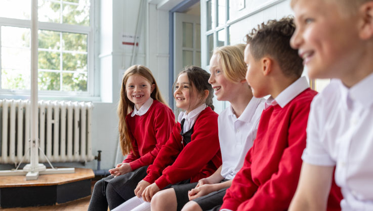A group of five school children sit in a row on a wooden bench inside a bright classroom. Most of the students are wearing red jumpers and white collared shirts, while some are in white polo shirts. They are all looking off-camera to the right with smiling, engaged expressions. Large windows in the background let in natural light.