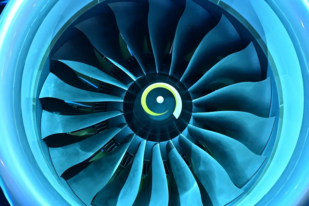 A close-up, brightly lit view of a jet engine turbine, showing the fan blades in shades of glowing electric blue, with a yellow and white swirl symbol in the center.