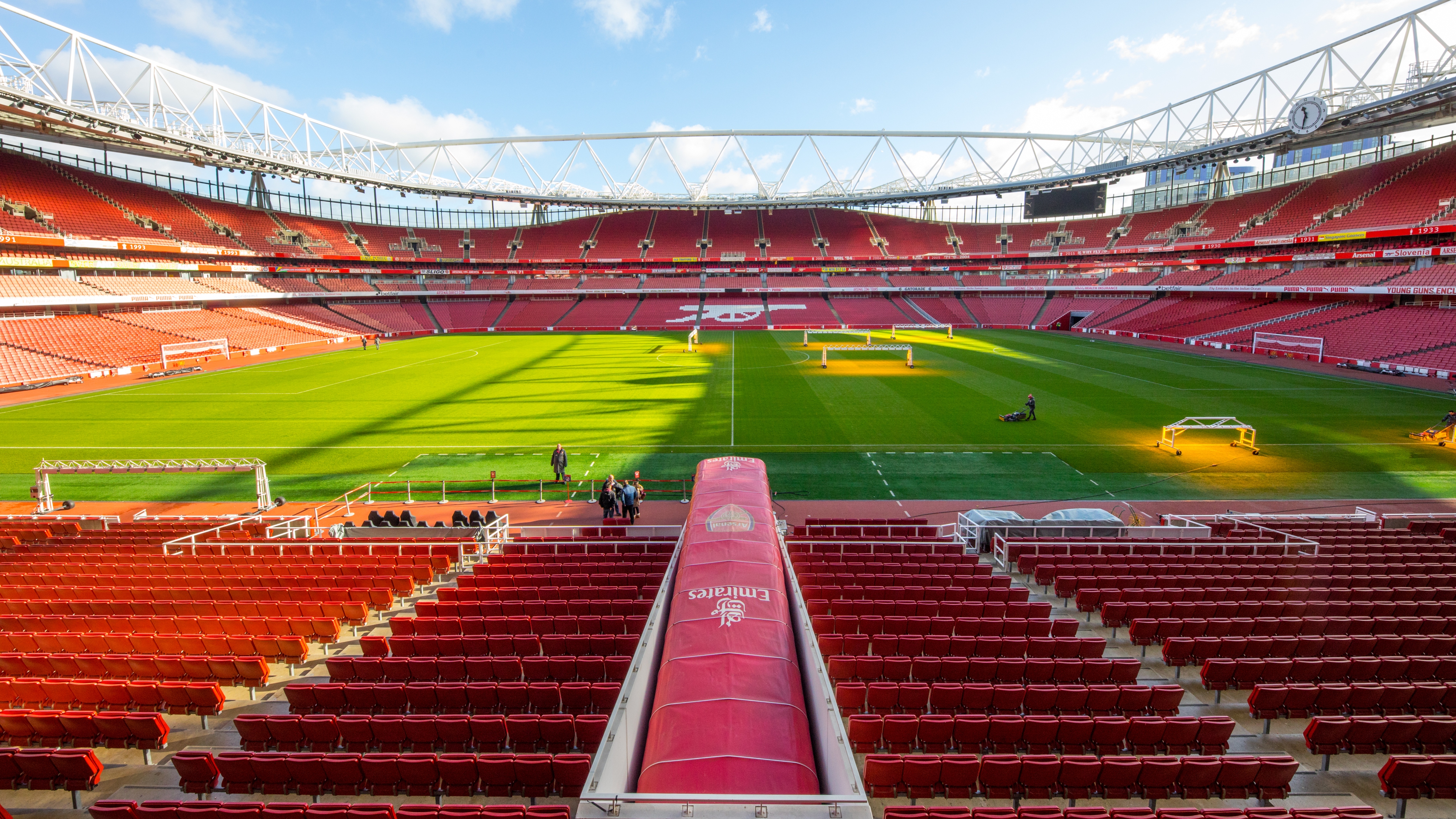 Wide view inside Emirates Stadium showing the football pitch and rows of red seating, with the players’ tunnel in the foreground and grounds staff working on the grass under bright daylight.
