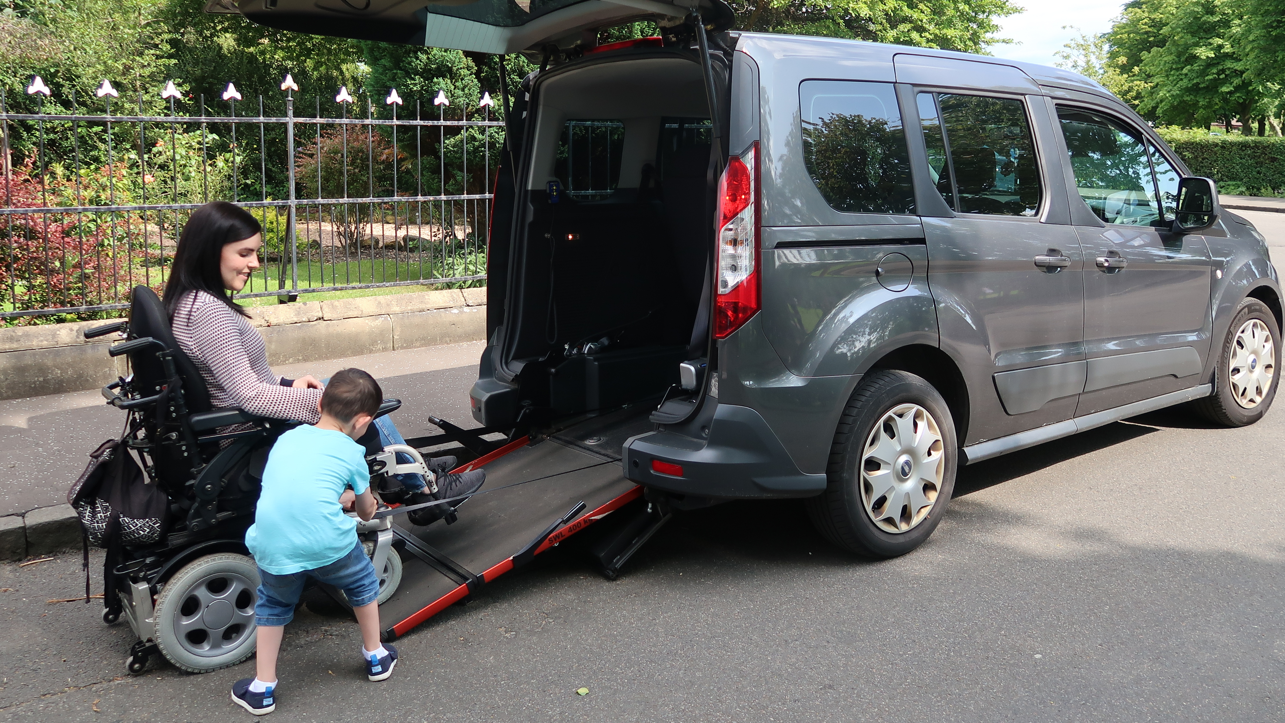 Emma Muldoon sits in her powered wheelchair at the base of a rear access ramp on a parked Wheelchair Accessible Vehicle (WAV) while a young child stands on the ramp beside her, with trees and a fence in the background.