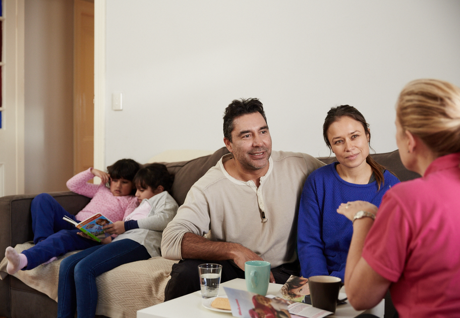 Family meeting with a support worker in their living room, while two children read together on the couch.