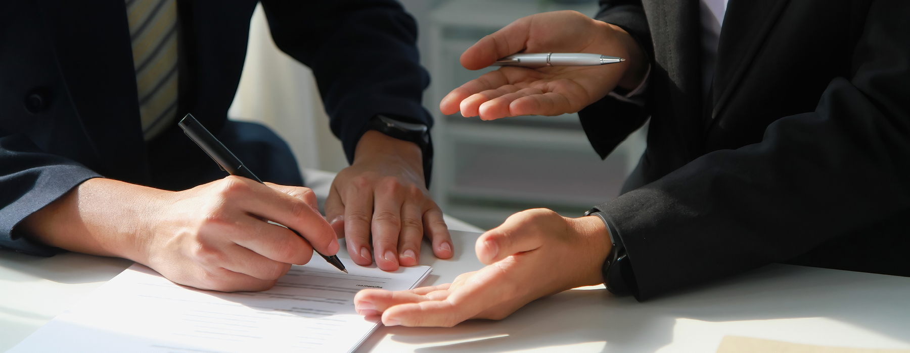 Two business people signing a contract. In view are their hands and a paper contract, with one person holding a pen over it as if they are about to sign.