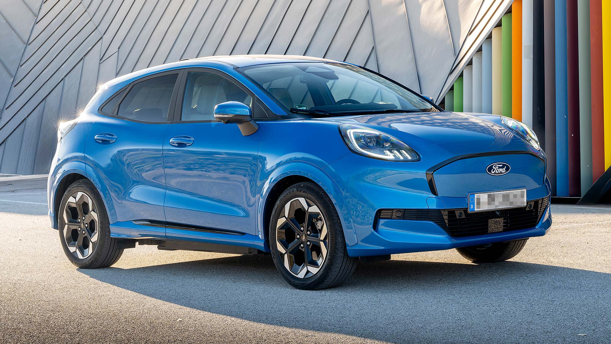 Blue Ford Puma Gen-E electric crossover parked in front of a modern architectural wall with colourful vertical panels.