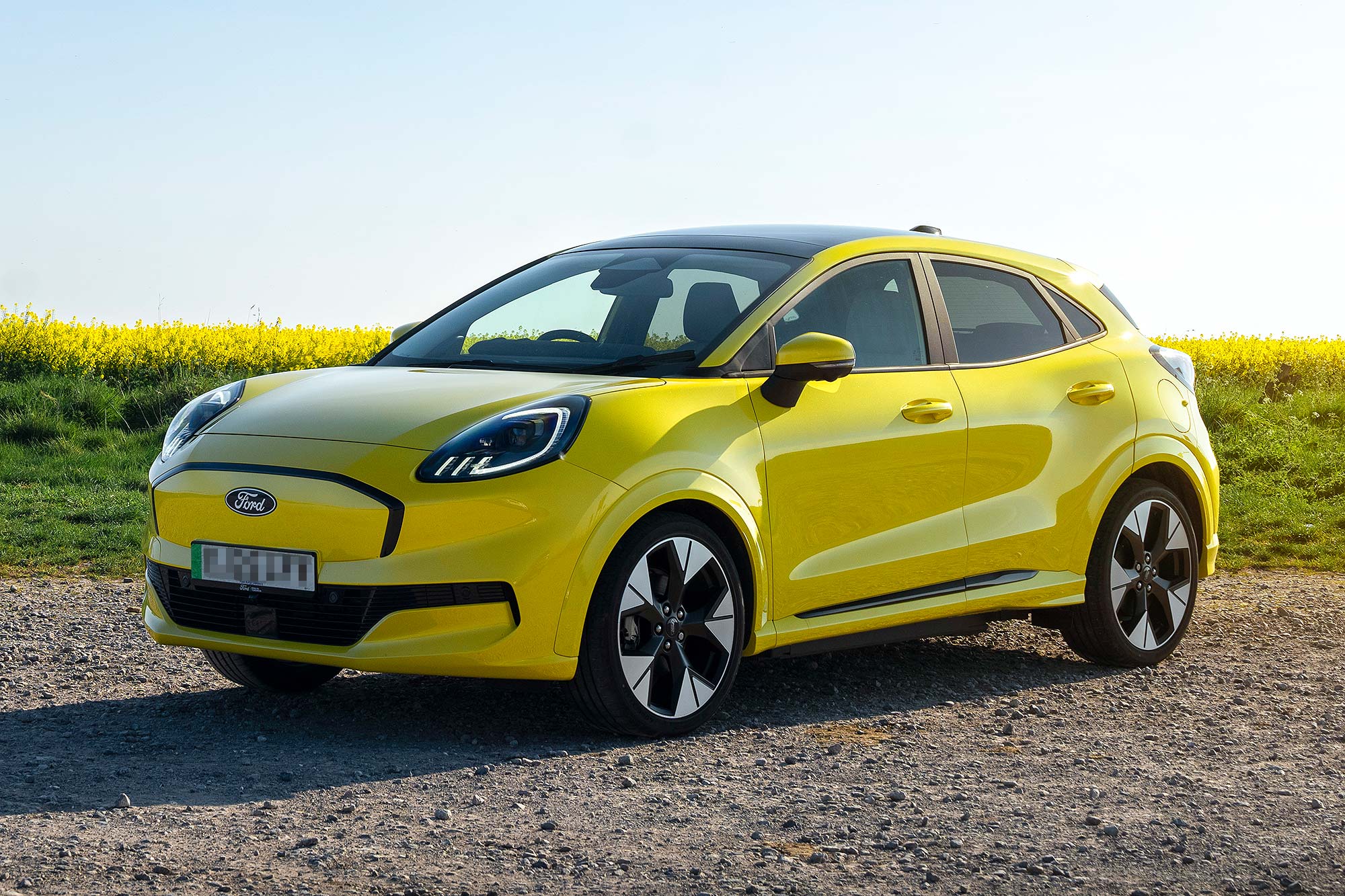 A yellow Ford Puma Gen-E electric crossover is parked on a rural gravel road beside a field of yellow flowers under bright sunlight.