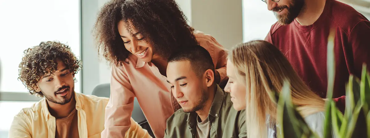 Five young adults gathered around a table in a bright office space, smiling and collaborating on a project, with one person pointing at something on the table."