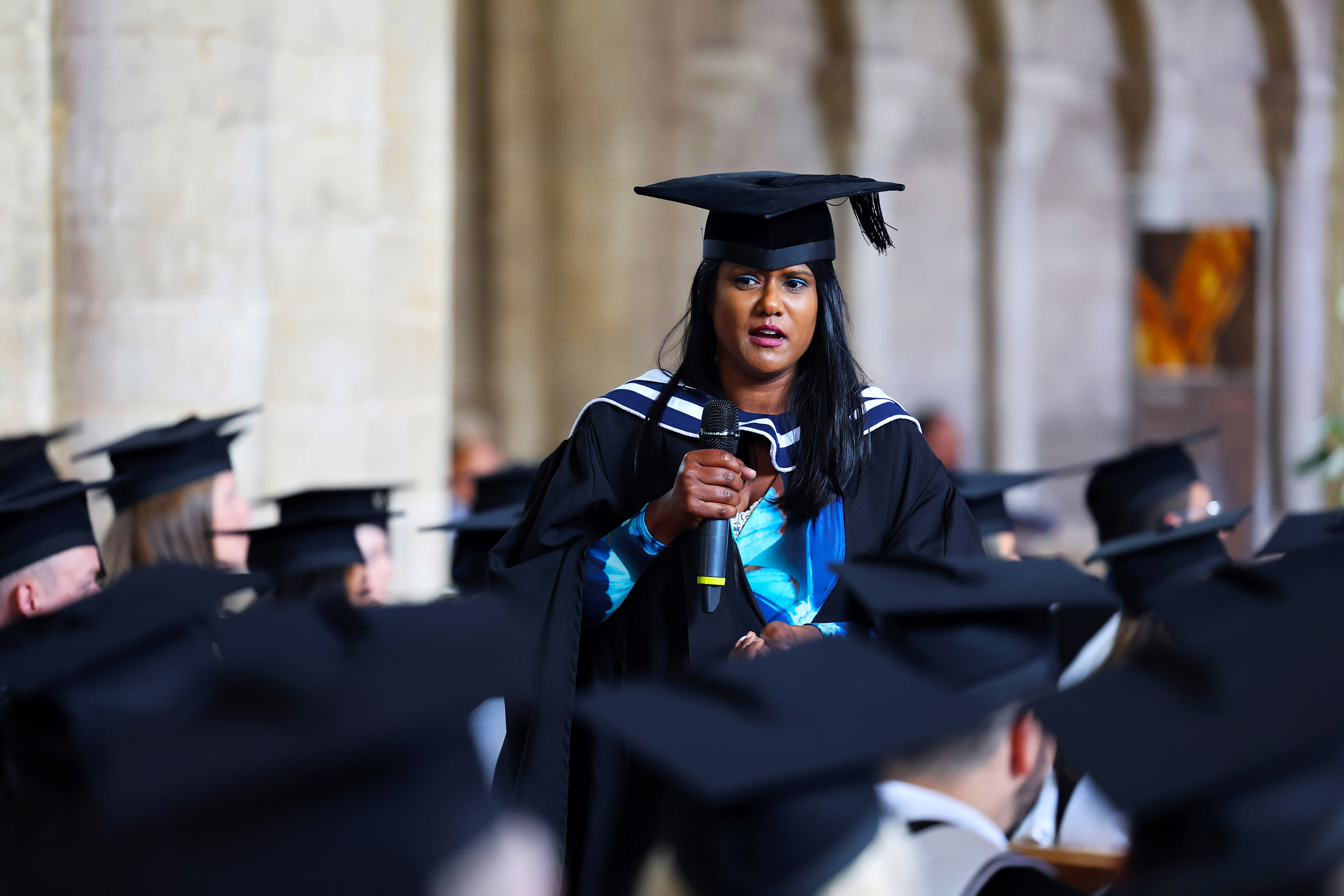 Graduate speaking into a microphone amidst the seated students