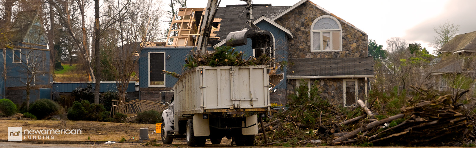 A partially damaged home made of wood and stone is surrounded by fallen trees. 