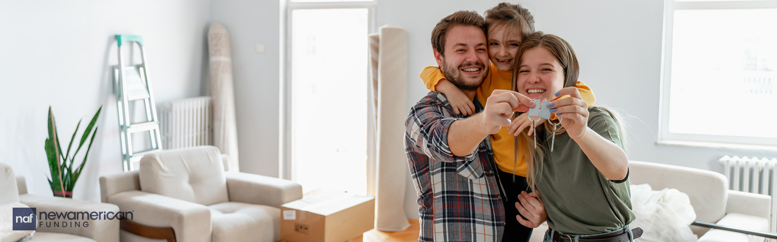 A young white man and woman stand smiling with a young girl behind them in a living room with boxes 