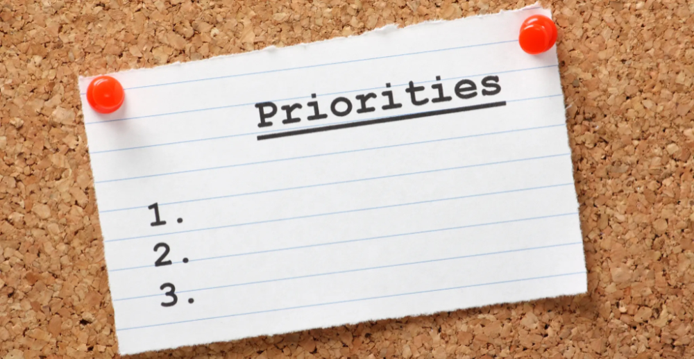 A corkboard with a piece of paper pinned to it saying "Priorities" and "1, 2, 3" on their own lines with space to write next to each number