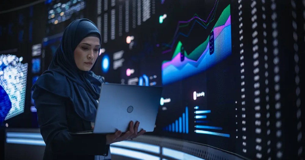 A woman wearing a black hijab standing in front of a curved screen wall showing data charts is holding a laptop