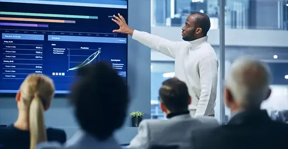 A man standing next to, and gesturing his hand to, a large screen showing data. In the foreground are the backs of four peoples heads, looking at the screen