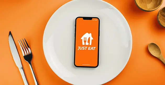 A table setting with a phone displaying the opening screen of the Just Eat app on a white dinner plate with a knife and fork on the left and a wooden spoon on the right