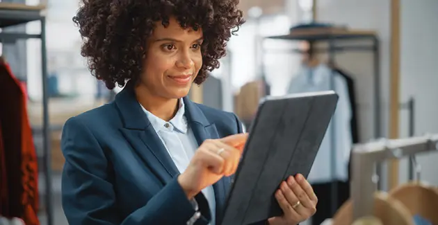 A woman wering a suit with dark brown curly hair holding an iPad, in the background are suits on hangers and rails