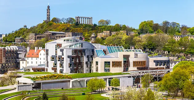 A photo of the Scottish Parliament Building in Holyrood, Edinburgh. It's a bright day with no clouds and lots of green trees are shown with the hill in the background