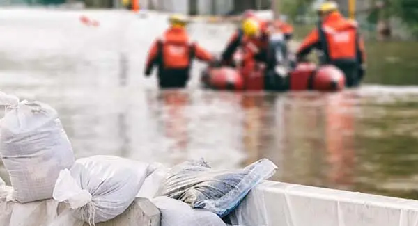 A blurry image of a flooded area with men in rescue gear walking alongside a boat, the water is up to their hips. In the foreground is sandbags 