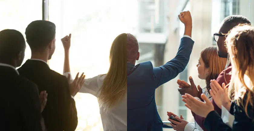 People on the left and right clapping and looking towards the middle back with a person with their back to the camera with fists raised in celebration. The left side of the person is a woman wearing a white shirt with long straight blonde hair and the right side of the person is a short haired man wearing a suit jacket. 