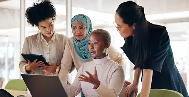 Three women stood over another woman sat at a laptop, they are in an office and wearing business attire