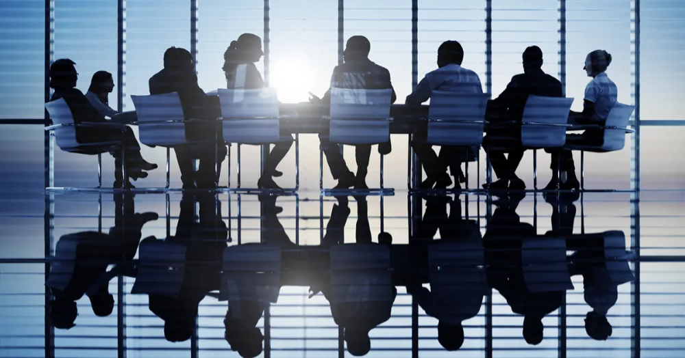 An image of a conference table with eight people sat around silhouetted against a large window and also the whole image reflected on the polished floor
