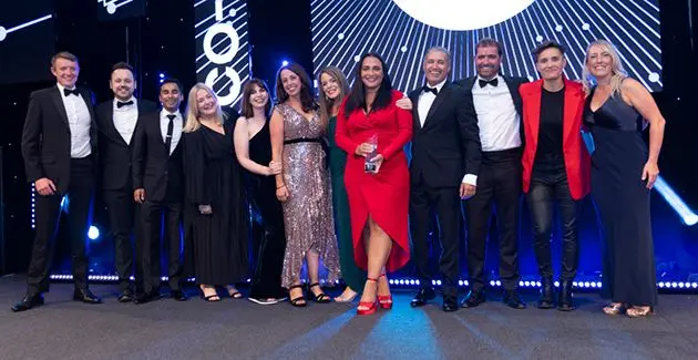 People stood in a line facing the camera, at an awards ceremony, smiling, wearing formal wear, arms around each other