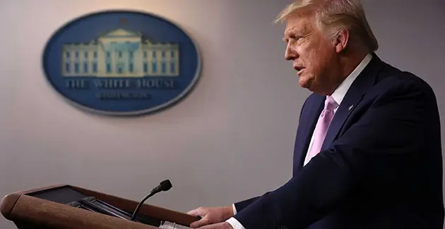 A side on photo of Donald Trump at a podium, wearing a suit, with The White House oval plaque on the wall in the background