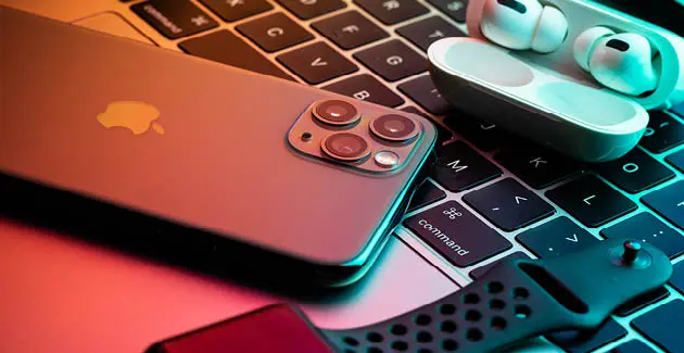 A close up photo of an iPhone face down on top of an apple laptop keyboard, also on top of the keyboard is a pair of earbuds in their case, open. As well as an apple watch 
