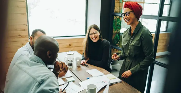 A meeting space with 4 people around a table with papers on, one of the people is a woman standing, the rest are sat down
