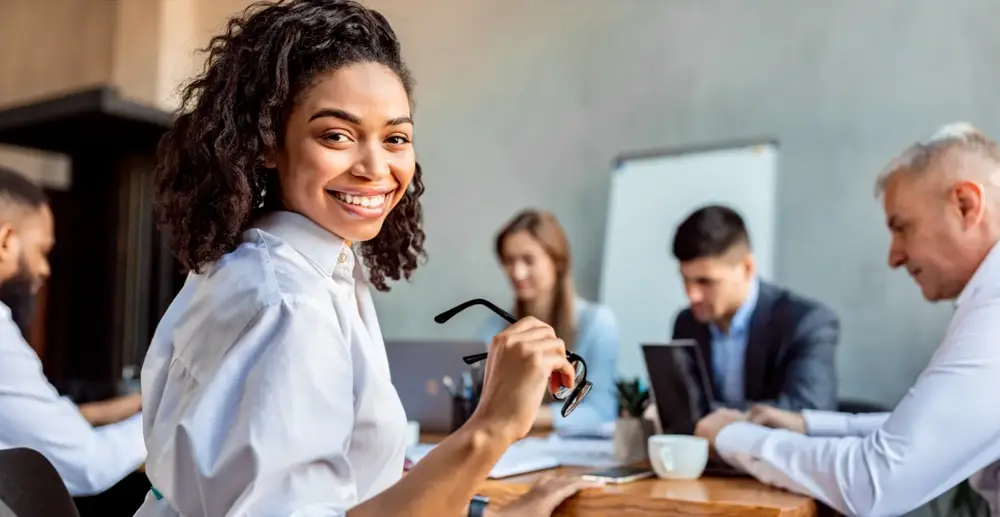 Woman turned to face the camera, smiling and holding a pair of glasses, behind her, slightly out of focus, sits four people sitting around a meeting room table. Everyone is wearing business attire