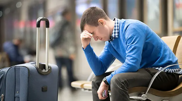 A man wearing a bright blue jumper sat on a public bench with his head in his hand looking sad, a suitcase is stood in front of him