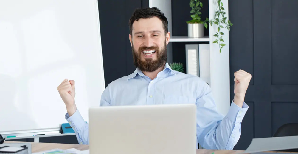 A man sat at a desk behind an open laptop celebrating with his arms up in fists and smiling
