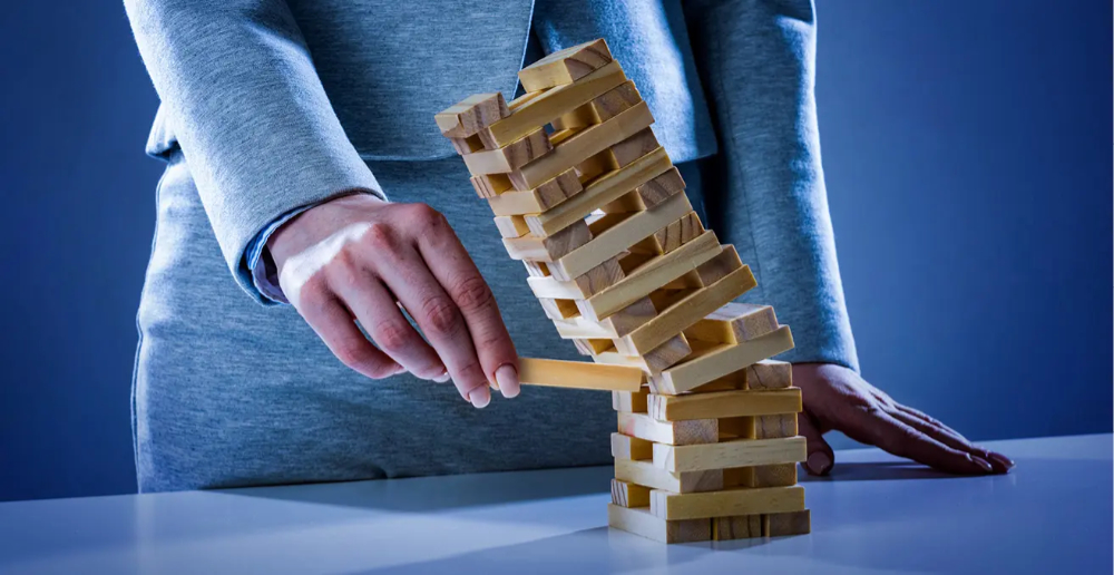 Jenga tower about to fall over as a womans hand is pulling a block out from below the middle point 