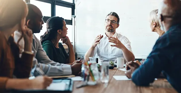 People sat around a conference table with a large whiteboard in the background, they're all looking at a man seated at the head of the table