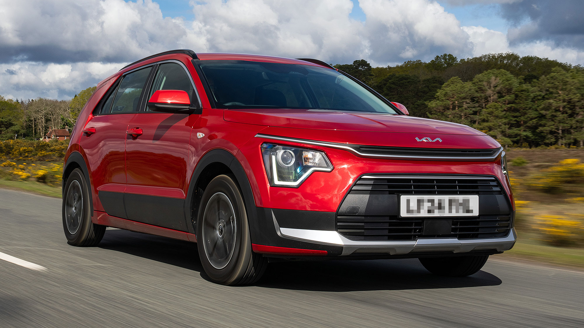 A red Kia Niro driving on a countryside road, with a distinctive angular headlight design and a sporty stance.