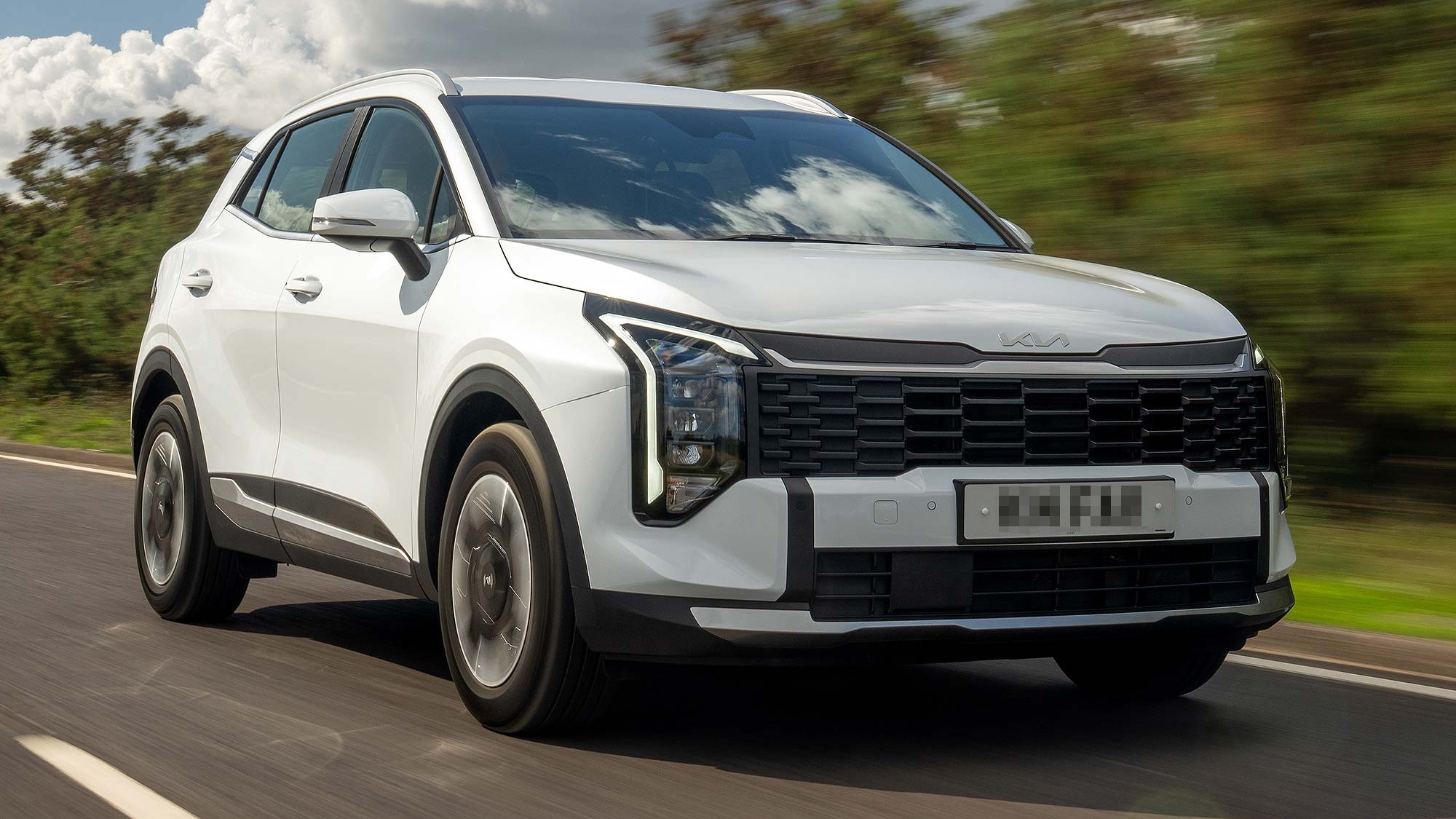 A white Kia Sportage SUV driving at speed on a road with blurred greenery, front three quarter view, available on the Motability Scheme.