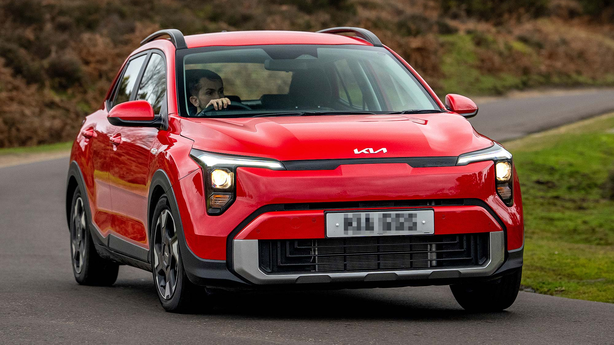 Red Kia Stonic compact SUV driving on a country road, shown from the front with the headlights on.