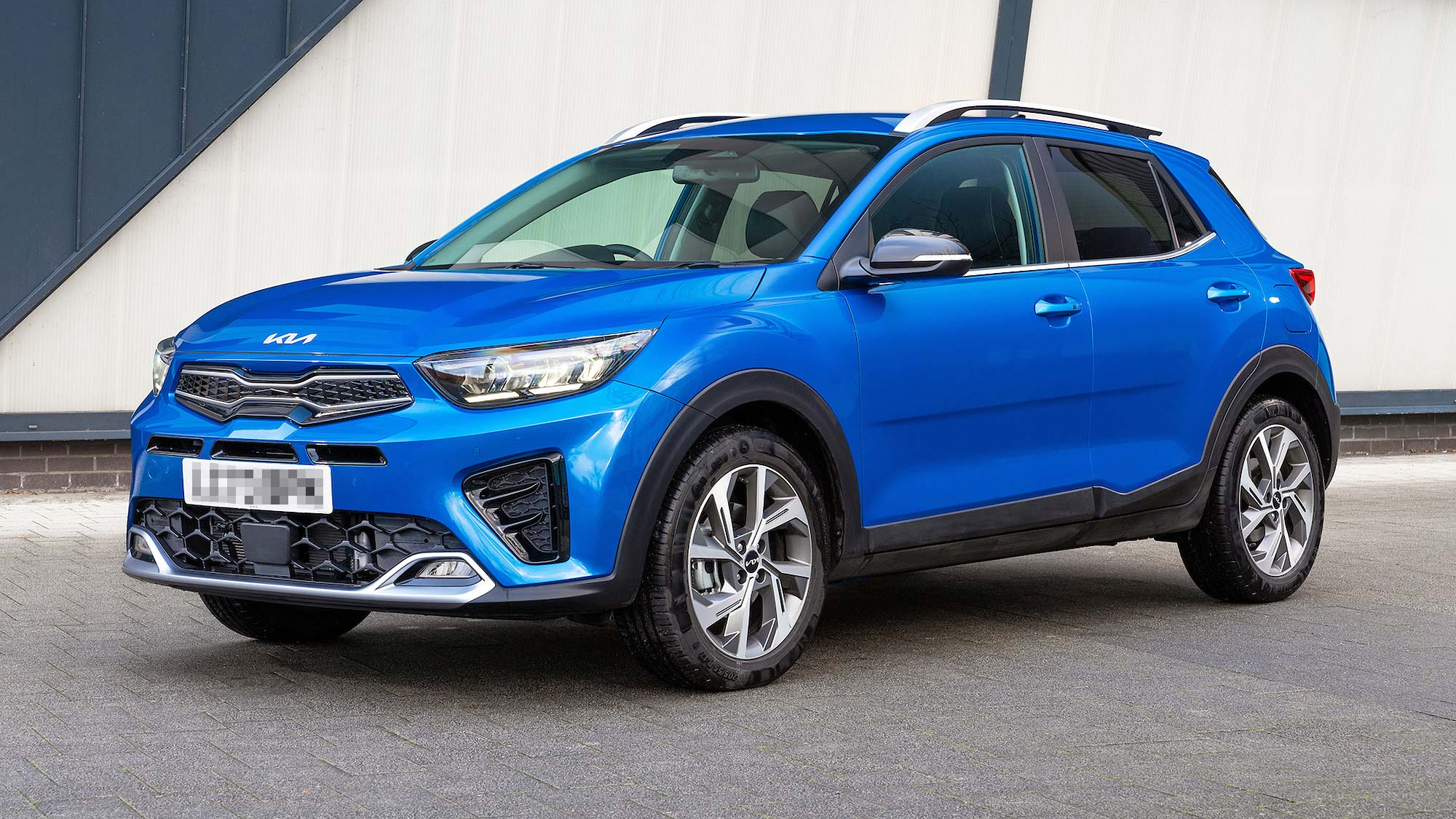 A blue Kia Stonic small SUV is parked on a paved surface in front of a building with grey and white walls.