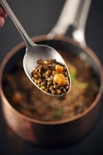 Spoon filled with cooked lentils in front of wooden bowl.