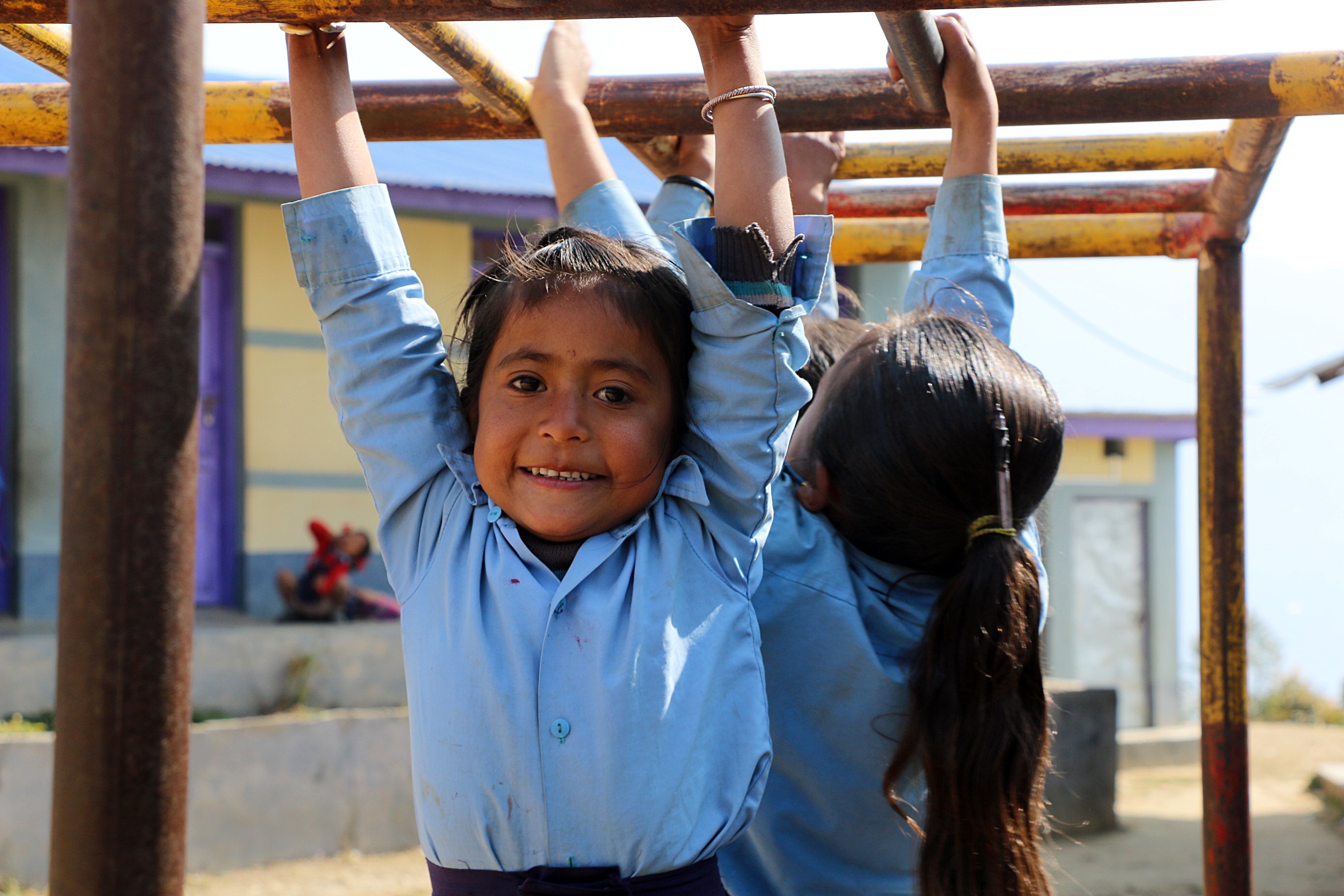 A young girl in a blue school uniform smiles brightly while hanging from yellow monkey bars on a sunny playground.