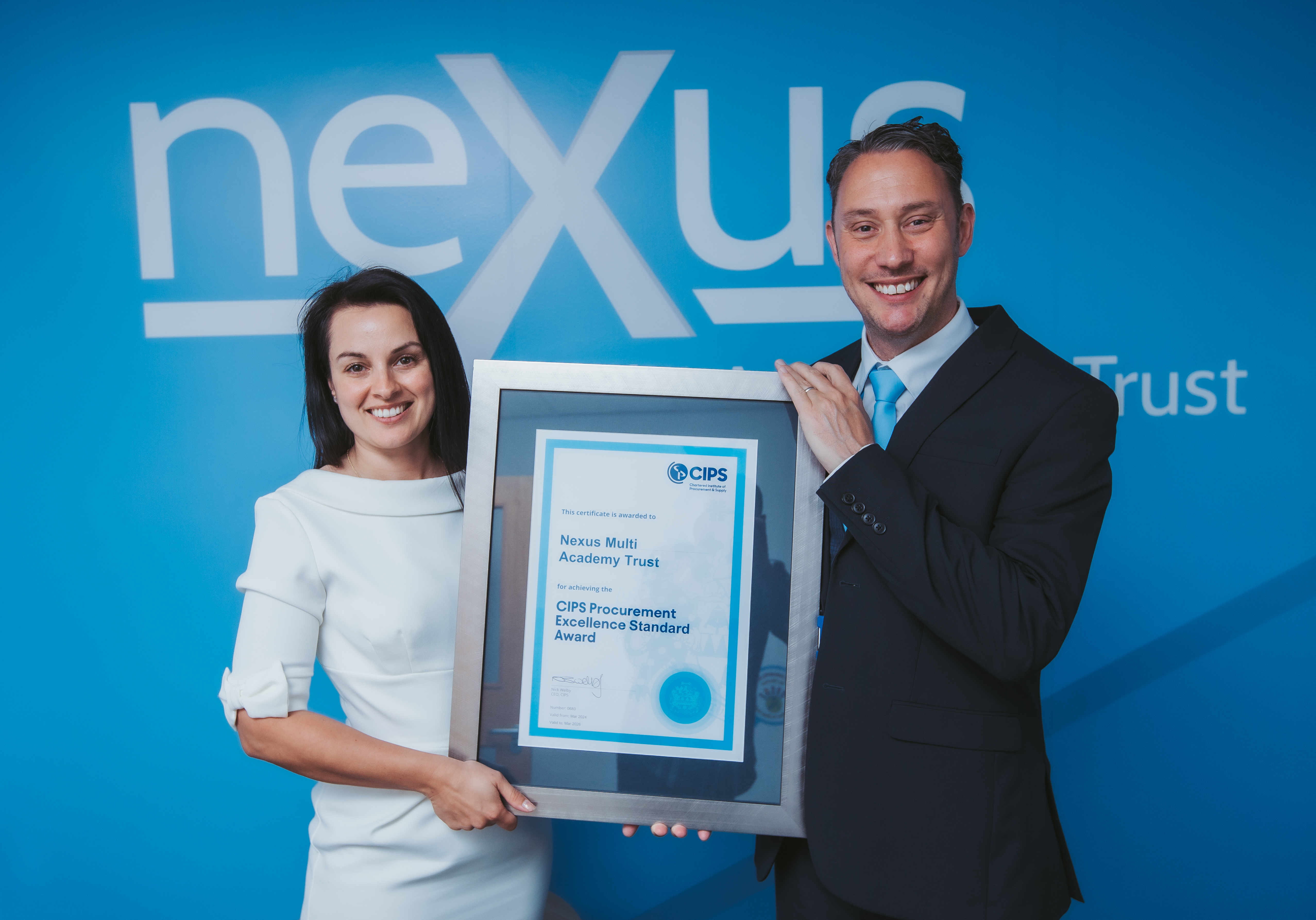 Smiling man and woman holding a frame with a CIPS Procurement Excellence Standard Award. In the background the Nexus logo is visible