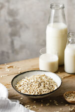Oats in a ceramic bowl and oat milk bottles and glass on table. 