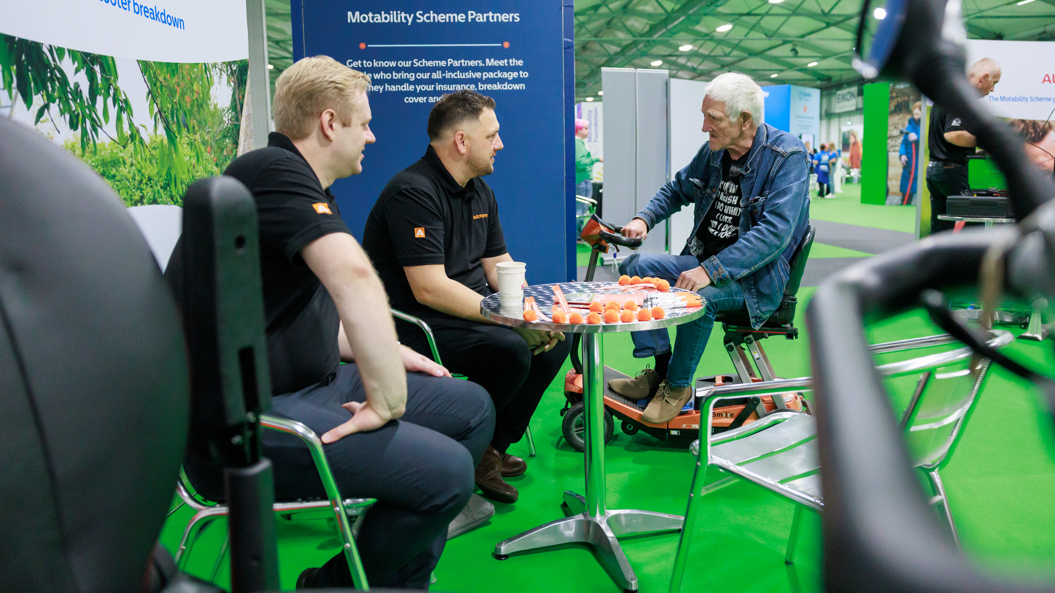 An older man seated on a mobility scooter talks with two Motability Scheme partners at a small table inside the exhibition hall.